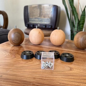 Op de afbeelding: Vier houten meubelfoten met een donkerhouten afwerking en een lichte houten afwerking. De poten zijn rond en hebben een schroef in het midden. Er zijn ook vier zwarte kunststof meubelfoten en een klein bakje met schroeven.