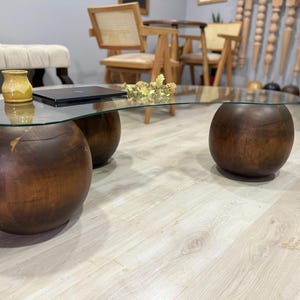 May include: A glass-top coffee table with a unique curved design. The table is supported by three large, round wooden legs. The table is made of dark wood and has a natural finish.