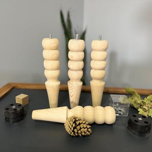 May include: A set of unfinished wooden furniture legs with a turned design, alongside hardware and a pine cone. The legs are light-colored and have a stacked, rounded appearance. Accessories include black mounting plates and a small wooden block.