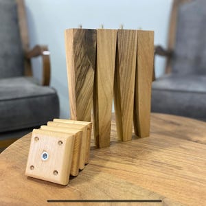 Könnte beinhalten: Satz von sechs Holzbeinen für Möbel, vier lang und zwei kurz. Die Beine sind aus hellbraunem Holz mit einer natürlichen Oberfläche. Die kurzen Beine haben eine quadratische Form und sind übereinander gestapelt. Die langen Beine sind spitz zulaufend und haben eine leicht abgerundete Form.