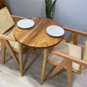Op de afbeelding: Twee houten stoelen met gevlochten rieten zittingen en rugleuningen, die tegenover een ronde houten tafel staan met twee witte borden. De tafel en stoelen zijn in een lichte houtkleur.