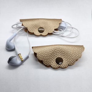 Könnte beinhalten: Zwei goldfarbene Leder-Kopfhörerhalter mit gewellten Kanten und einem Knopfverschluss. Die Halter sind leer, aber einer hat weiße Kopfhörer darum gewickelt.
