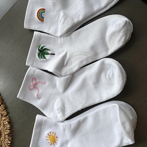 May include: Four pairs of white ankle socks with embroidered designs. One sock features a rainbow, another a palm tree, a third a pink bow, and the last a yellow sun. The socks are arranged on a gray surface.