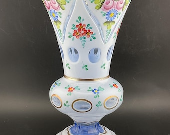 Op de afbeelding: Een witte glazen vaas met een blauwe en gouden rand. De vaas is versierd met handgeschilderde bloemen in tinten roze, rood, blauw en groen.
