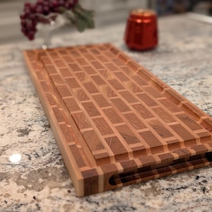 Puede incluir: Una tabla de cortar rectangular de madera con un patrón similar a ladrillos. La tabla es de madera marrón clara y oscura, con una ranura para jugos y huecos para las asas. Una lata roja y un jarrón con uvas están en el fondo.