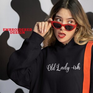 May include: Black hooded sweatshirt with the text "Old Lady -ish" embroidered in white on the front. The sweatshirt is being worn by a person with red sunglasses and a red crossbody bag. The image includes the logo "Sweet Honey Bun Designs" in the bottom right corner.