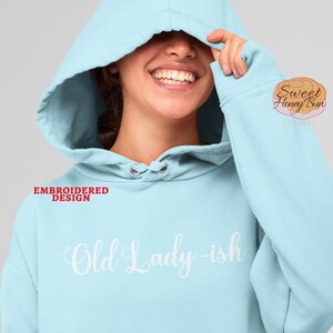 May include: A light blue hooded sweatshirt with the text "Old Lady-ish" embroidered in white on the front. The text "EMBROIDERED DESIGN" is in red on the left side of the sweatshirt.