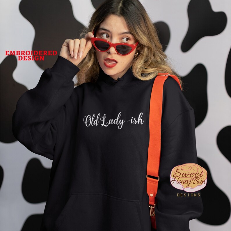 May include: Black hooded sweatshirt with the text "Old Lady -ish" embroidered in white on the front. The sweatshirt is being worn by a person with red sunglasses and a red crossbody bag. The image includes the logo "Sweet Honey Bun Designs" in the bottom right corner.