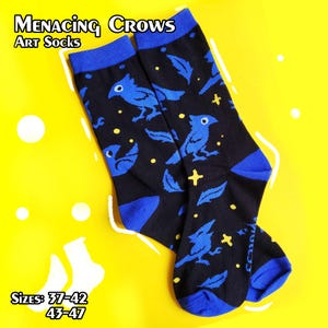 Op de afbeelding: Een paar zwarte kunstsokken met een blauw kraaien- en verenpatroon, blauwe manchetten en tenen. De sokken staan op een gele achtergrond. Tekst op de sokken luidt "Menacing Crows Art Socks". Maten 37-42 en 43-47 worden vermeld.