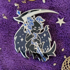 May include: A black and blue enamel pin depicting a small, winged creature holding a silver scythe. The creature has a dark blue head with a star-shaped pattern on its wings.