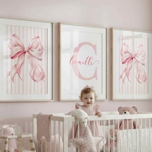May include: A nursery scene with a white crib, high chair, and decorative items. Three framed prints hang above the crib, featuring pink bows and the name "Camille." The room is painted in soft pink tones, creating a gentle and inviting atmosphere.