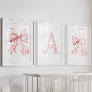 May include: Three framed wall art pieces. Two feature watercolor pink bows, and one displays the name "Avery" in a pink script font. The room includes a white crib, chairs, and a small table, creating a nursery theme.