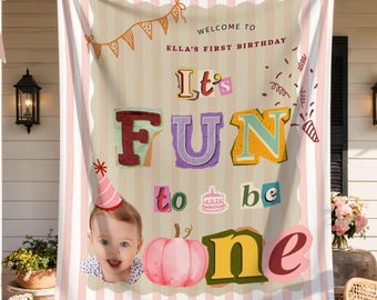 May include: A large 68 by 80 inch fabric banner with pink and white vertical stripes. The center features a beige panel with the text "WELCOME TO ELLA'S FIRST BIRTHDAY" and "It's FUN to be one," a photo of a baby, a pink pumpkin, and a cake.