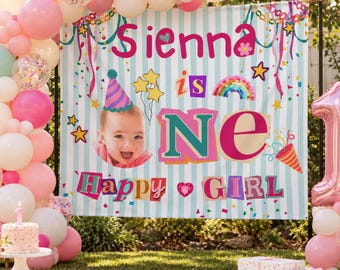 May include: A birthday backdrop featuring light blue and white vertical stripes, the text Sienna is ONE Happy GIRL, a photo of a baby in a party hat, a rainbow, and confetti. A large pink balloon shaped like the number one stands to the right.