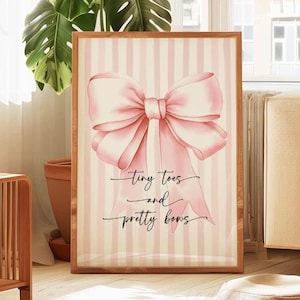May include: A framed print featuring a large pink bow illustration on a striped background. The text "tiny toes and pretty bows" is written in cursive below the bow. The frame is a light brown color.