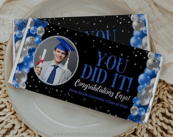 May include: A black and silver graduation chocolate bar with blue balloons and confetti. The bar has a photo of a graduate wearing a cap and gown and holding a diploma. The text on the bar reads "You Did It! Congratulations Enzo! Stony Brook University - Class of 202X".