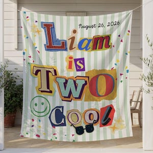 May include: A white and light green striped blanket featuring the text Liam is TWO Cool and August 26, 2026. The design includes colorful cutout-style letters, a green smiley face, black sunglasses, and scattered confetti shapes in various colors.