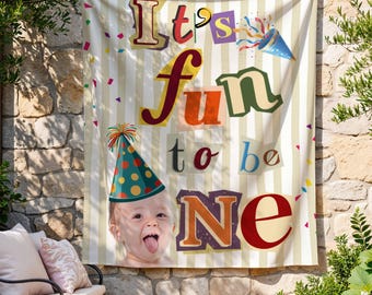 May include: A rectangular fabric wall hanging with a beige and white vertical striped background. The text reads It's fun to be NE in colorful block letters. A party hat and confetti are printed alongside a photo of a baby sticking out their tongue.