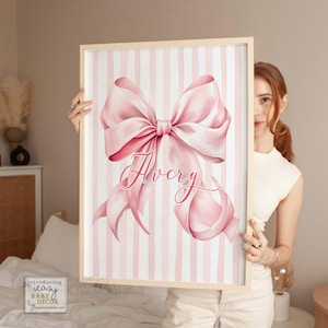 May include: A framed print featuring a large pink bow with the name "Avery" in cursive script. The background has vertical pink and white stripes. The frame is made of light-colored wood. The print is a decorative item.