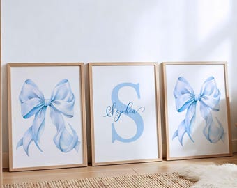 May include: Three framed wall art prints with light wooden frames. The two outer prints feature a large, light blue ribbon bow. The center print displays a large light blue letter S with the name Sophia written in blue cursive script.