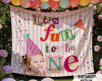 May include: A rectangular fabric backdrop featuring a light pink and white vertical striped pattern. The text "It's fun to be ONE" is printed in colorful, blocky letters. A pink and teal party hat and confetti graphics are visible. Size: 50"x60".