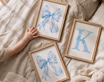 May include: Three rectangular wooden frames containing wall art prints. Two prints feature a light blue ribbon bow illustration on a white background. The third print displays a large light blue letter K with the name Katy written in blue script.