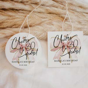 May include: Two round and rectangular white gift tags with pink and gold watercolor bow designs. The text on the tags reads "Cheers to 30 years!" and "Mariela's Birthday 10 | 18 | 2022".