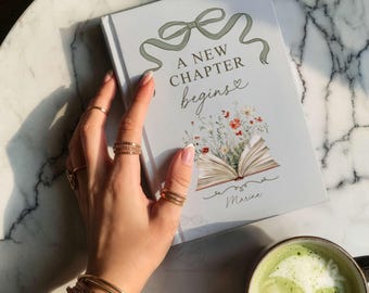 May include: A white hardcover book featuring a sage green ribbon illustration at the top, the text A NEW CHAPTER begins, and a colorful floral graphic emerging from an open book. The name Marina is printed in cursive at the bottom of the cover.