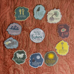 May include: A set of 12 colorful stickers with inspirational quotes and illustrations. The stickers are circular and feature a variety of designs, including a butterfly, a key, a sun, and a moon. The stickers are arranged in a circle around a central sticker that reads "Letters to the (Re)atrix - Durga Alas".