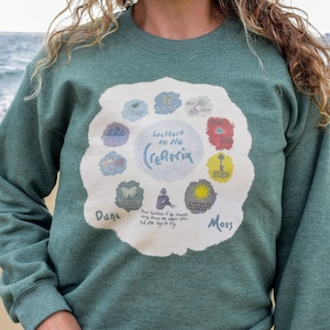 May include: A green sweatshirt with a white circle graphic featuring the text "lectures to the (re)matrix" and a series of colorful circles with images and text. The text "Dune" and "Moss" are visible at the bottom of the graphic.
