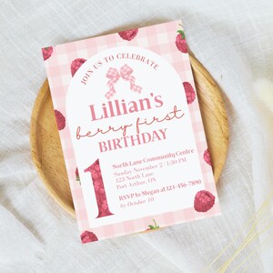 Puede incluir: Una invitación de cumpleaños rosa a cuadros con ilustraciones de frambuesas. La invitación dice "Lillian's berry first BIRTHDAY" con detalles del evento. El número "1" se muestra de forma destacada. La invitación está sobre un plato de madera.