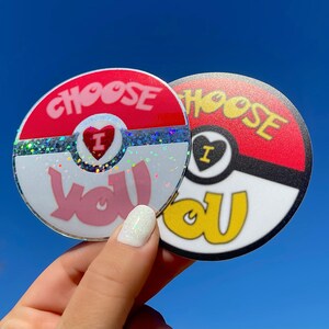 K&ouml;nnte beinhalten: Zwei runde Aufkleber mit einem roten, wei&szlig;en und schwarzen Design. Die Aufkleber sind beide Pok&eacute; Ball-thematisch und sagen "Choose You" in rosa und gelben Buchstaben. Ein Aufkleber hat Glitzer auf dem wei&szlig;en Teil des Pok&eacute; Balls.