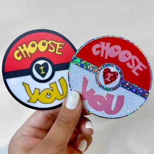 K&ouml;nnte beinhalten: Zwei runde Aufkleber mit einem roten und wei&szlig;en Pokeball-Design. Der Text "Choose You" ist in Pink und Gelb auf jedem Aufkleber geschrieben. Ein Aufkleber hat eine glitzernde Oberfl&auml;che.
