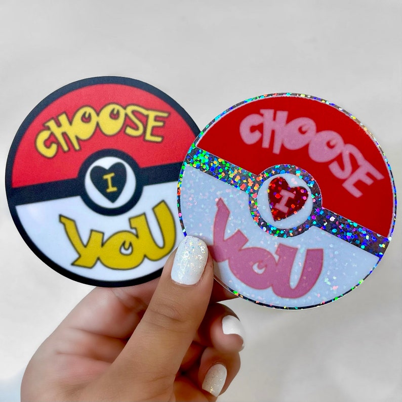 K&ouml;nnte beinhalten: Zwei runde Aufkleber mit einem roten und wei&szlig;en Pokeball-Design. Der Text "Choose You" ist in Pink und Gelb auf jedem Aufkleber geschrieben. Ein Aufkleber hat eine glitzernde Oberfl&auml;che.