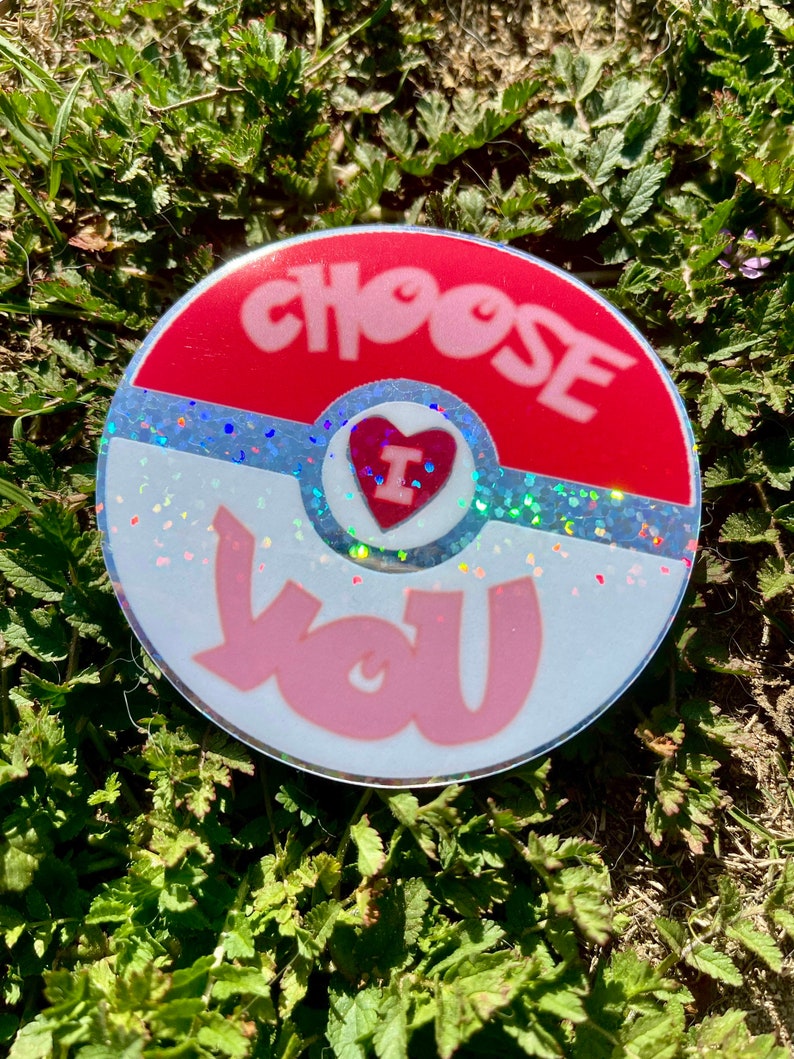 K&ouml;nnte beinhalten: Ein runder Aufkleber in Rot, Wei&szlig; und Blau mit Glitzer. Der Aufkleber tr&auml;gt die Worte "CHOOSE" und "YOU" in rosa Schrift. Ein rotes Herz mit einem wei&szlig;en "I" darin befindet sich in der Mitte des Aufklebers.