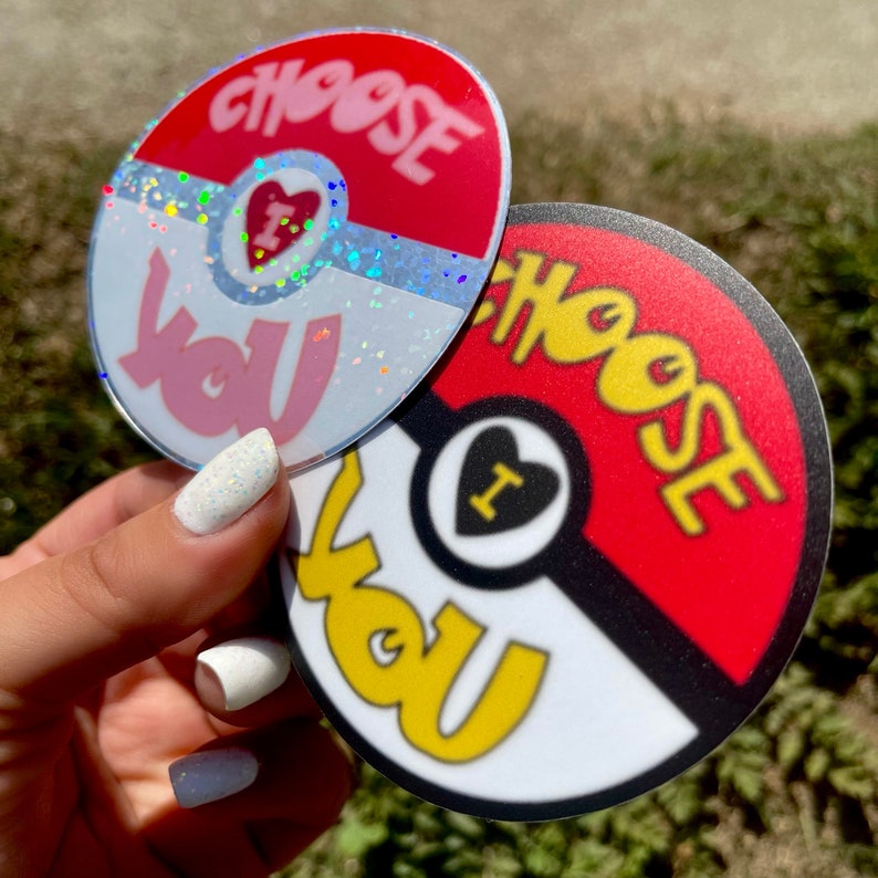 K&ouml;nnte beinhalten: Zwei runde Aufkleber mit einem roten, wei&szlig;en und schwarzen Pokeball-Design. Der Text "Choose You" ist in Gelb auf beiden Aufklebern geschrieben. Ein Aufkleber hat ein pinkfarbenes Herz mit dem Buchstaben "I" darin und Glitzer. Der andere Aufkleber hat ein schwarzes Herz mit dem Buchstaben "I" darin.
