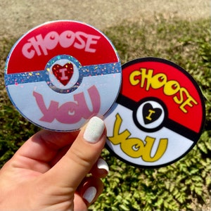 K&ouml;nnte beinhalten: Zwei runde Aufkleber mit einem roten, wei&szlig;en und schwarzen Pokeball-Design. Der Text "Choose You" ist in einer stilisierten Schrift auf beiden Aufklebern geschrieben. Ein Aufkleber hat ein rosa Herz mit dem Buchstaben "I" darin, und der andere hat ein schwarzes Herz mit dem Buchstaben "I" darin.