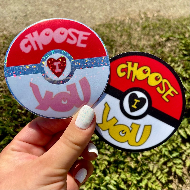 K&ouml;nnte beinhalten: Zwei runde Aufkleber mit einem roten, wei&szlig;en und schwarzen Pokeball-Design. Der Text "Choose You" ist in einer stilisierten Schrift auf beiden Aufklebern geschrieben. Ein Aufkleber hat ein rosa Herz mit dem Buchstaben "I" darin, und der andere hat ein schwarzes Herz mit dem Buchstaben "I" darin.