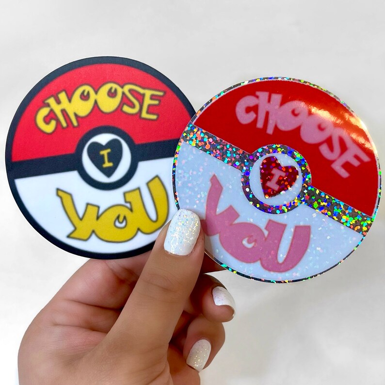 K&ouml;nnte beinhalten: Zwei runde Aufkleber mit einem roten und wei&szlig;en Pokeball-Design. Der Text "Choose You" ist in Gelb und Pink auf jedem Aufkleber geschrieben. Ein Aufkleber hat eine glitzernde Oberfl&auml;che.