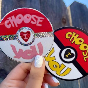 K&ouml;nnte beinhalten: Zwei runde Aufkleber mit einem roten, wei&szlig;en und schwarzen Pokeball-Design. Der obere Aufkleber sagt "Choose You" in rosa Buchstaben mit einem roten Herzen, in dem "I" steht. Der untere Aufkleber sagt "Choose You" in gelben Buchstaben.