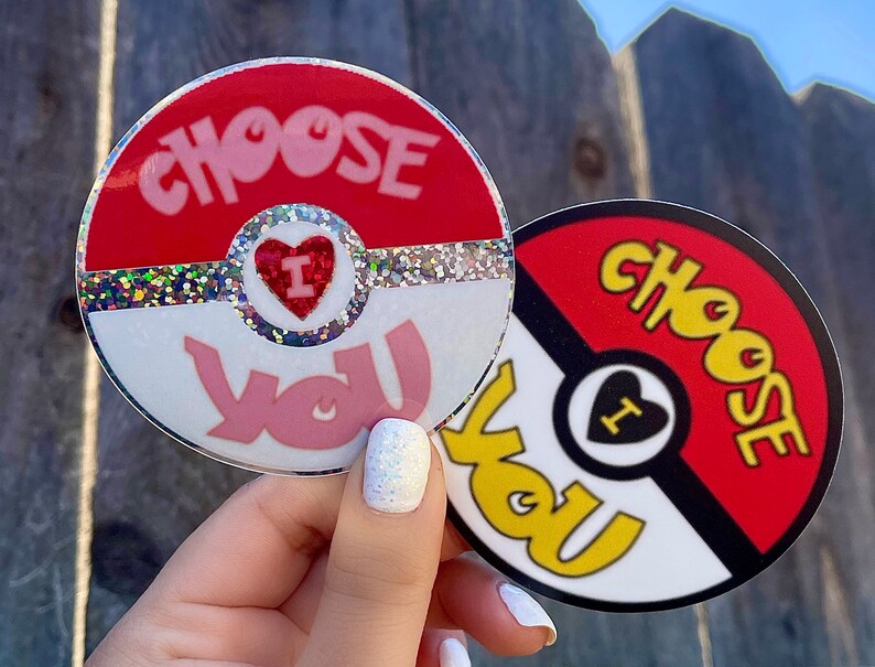 K&ouml;nnte beinhalten: Zwei runde Aufkleber mit einem roten, wei&szlig;en und schwarzen Pokeball-Design. Der obere Aufkleber sagt "Choose You" in rosa Buchstaben mit einem roten Herzen, in dem "I" steht. Der untere Aufkleber sagt "Choose You" in gelben Buchstaben.