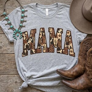 May include: A heather grey short-sleeve t-shirt featuring the word MAMA in a block font with a brown and tan cowhide, leopard print, and cactus pattern. The shirt is displayed with a turquoise squash blossom necklace, a brown hat, and boots.