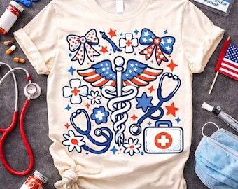 Op de afbeelding: Crèmekleurig T-shirt met een medisch caduceus-ontwerp met elementen van de Amerikaanse vlag. Het ontwerp bevat rode, witte en blauwe sterren, strikken, bloemen en stethoscopen. Een EHBO-koffer en de tekst "Registered Nurse" zijn ook zichtbaar.