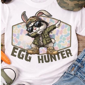 May include: White t-shirt with a cartoon rabbit wearing sunglasses and a camouflage hat, dabbing. The shirt has the text "EGG HUNTER" and is surrounded by a colorful pattern. Sunglasses, a hat, and sneakers are also visible.