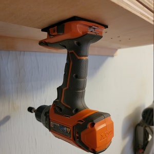 May include: An orange and black cordless drill is mounted upside down on a wooden shelf. The drill has a black handle and a black and orange body. The drill is labelled "X-Drive".