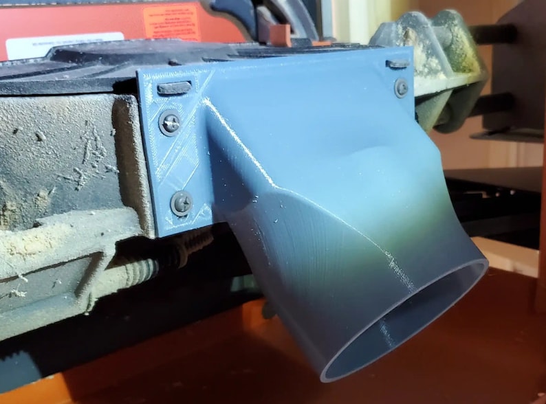 Pode incluir: Um adaptador de coleta de p&oacute; cinza impresso em 3D com uma abertura redonda e quatro parafusos. O adaptador est&aacute; preso a uma m&aacute;quina preta e laranja.