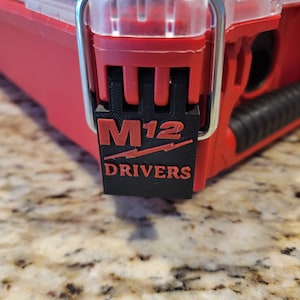 Può includere: Una cassetta degli attrezzi rossa e nera con un fermo in metallo e un'etichetta nera che dice "M12 DRIVERS".