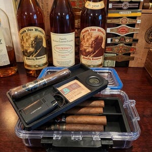 May include: Three bottles of Pappy Van Winkle bourbon, a clear plastic cigar travel case with a black interior tray, a Vertigo lighter, a Boveda 69% humidity pack, a digital hygrometer, and several cigars. The background features various cigar bands.