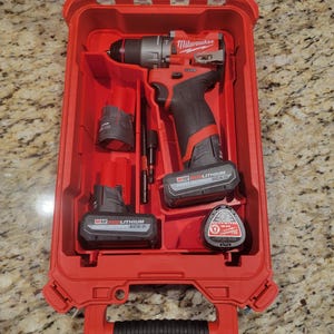Peut inclure: Une mallette à outils Milwaukee rouge avec une poignée noire. La mallette contient une perceuse sans fil rouge et noire, deux batteries rouge et noire et un porte-embouts noir.
