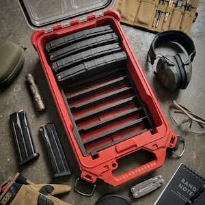 Könnte beinhalten: Eine rote und schwarze taktische Aufbewahrungsbox mit transparentem Deckel, gefüllt mit schwarzen Magazinen. Das Bild zeigt auch eine beige Werkzeugrolle, schwarzen Gehörschutz, eine Schutzbrille, einen Notizblock und weiteres Zubehör.
